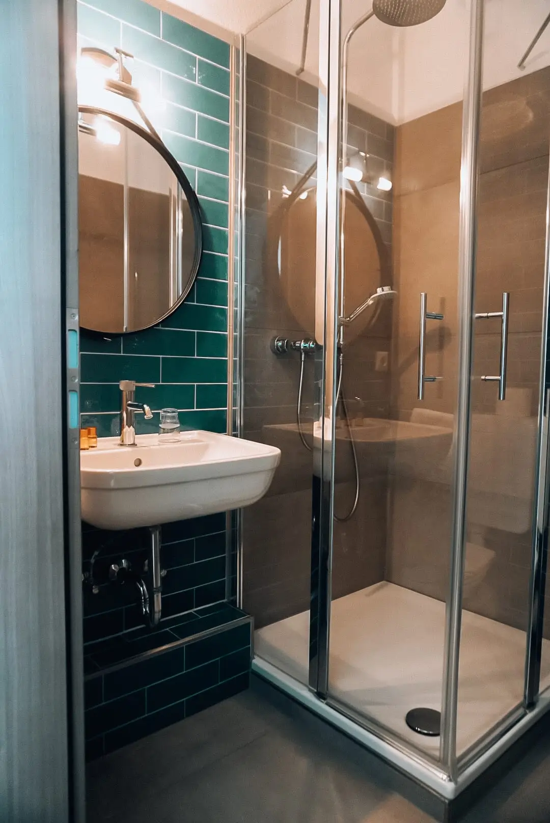 Badezimmer mit rundem Spiegel, grünen Wandfliesen und verglaster Eckdusche mit Handbrause