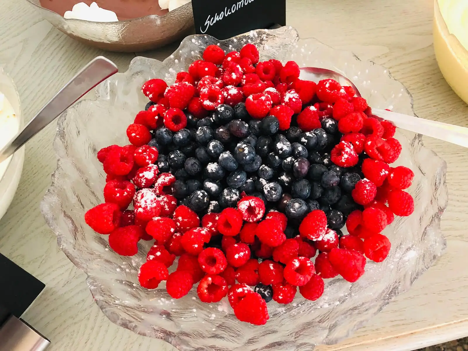 Glasschale mit Himbeeren und Heidelbeeren unter Puderzucker, daneben Schild für Schokomousse
