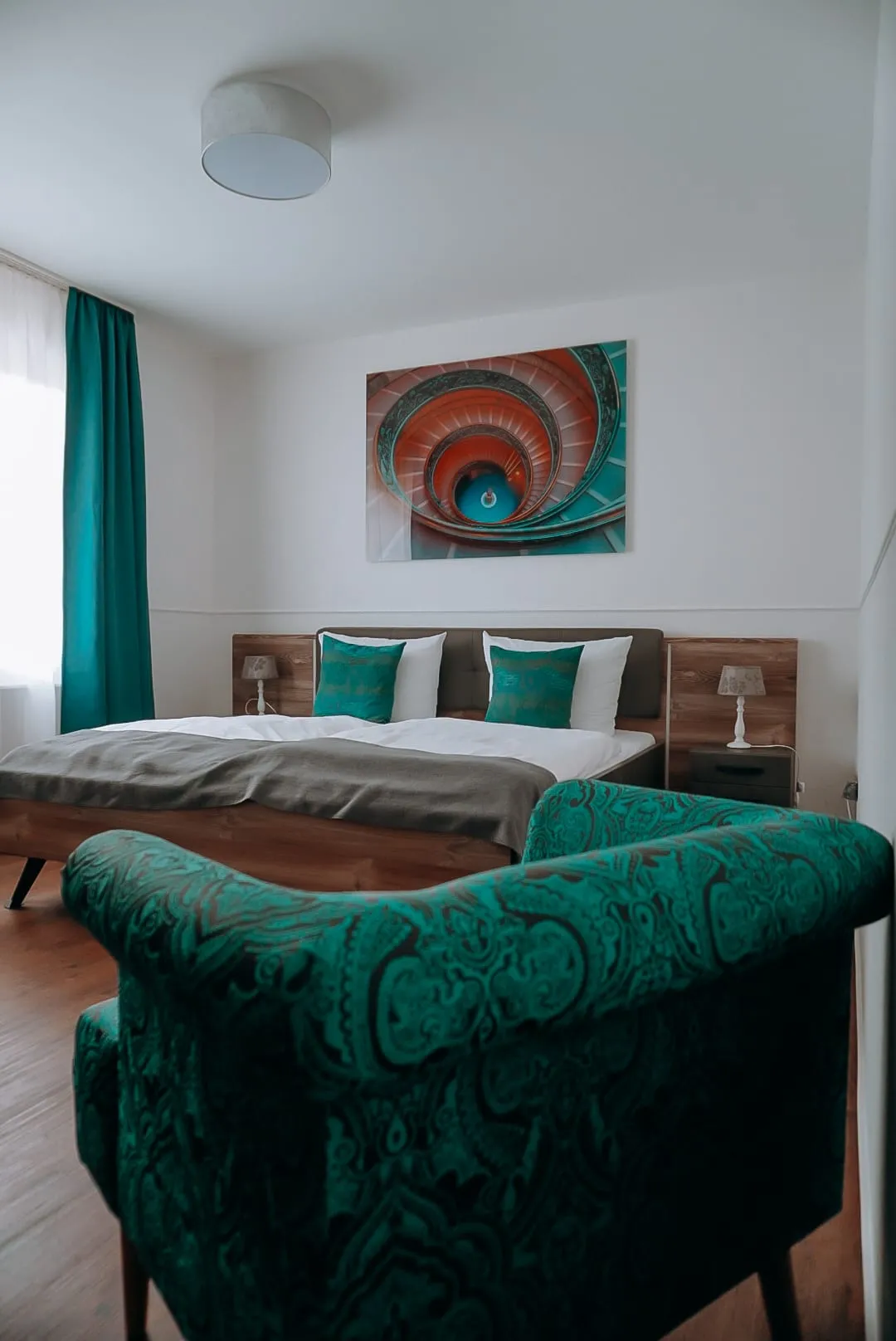 Doppelzimmer de luxe mit Holzbett, türkisen Kissen, Wendeltreppenbild und grünem Ornamentsessel