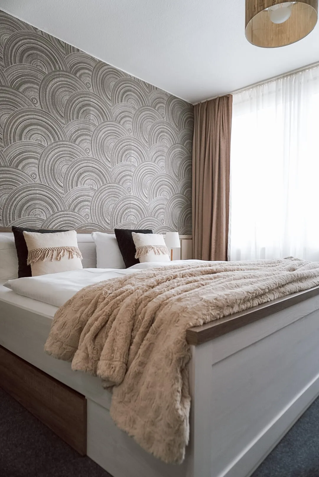 Doppelbett mit weißer Bettwäsche und beiger Felldecke vor gemusterter Kreistapete am Fenster
