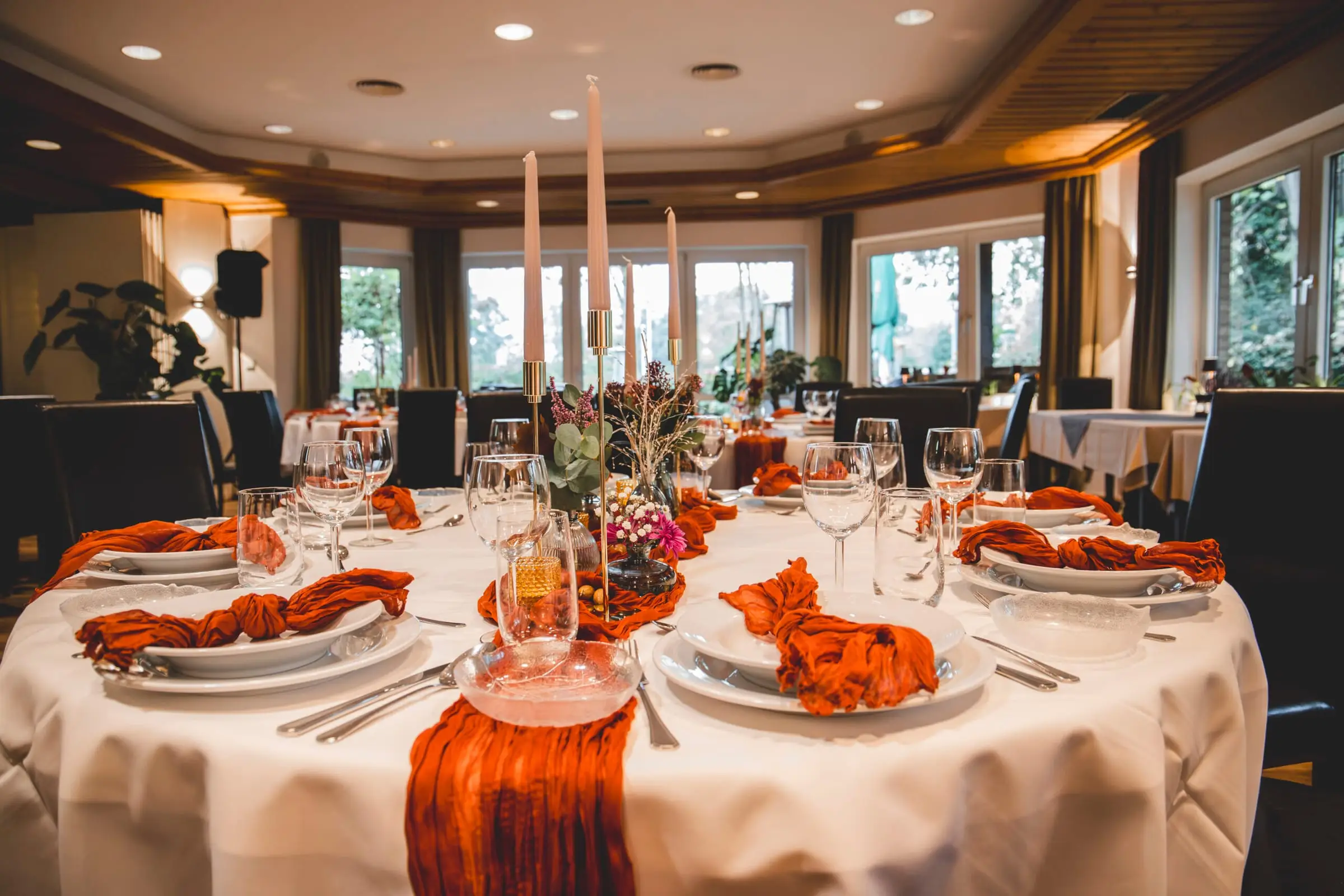 Runde Festtafel mit terrakottafarbenen Servietten, hohen Kerzen und Trockenblumen am Fenster