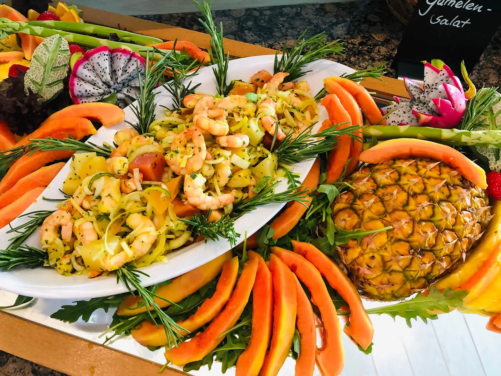 Garnelensalat auf ovaler Platte, umlegt mit Papayaspalten und Rosmarin, daneben ausgehöhlte Ananas