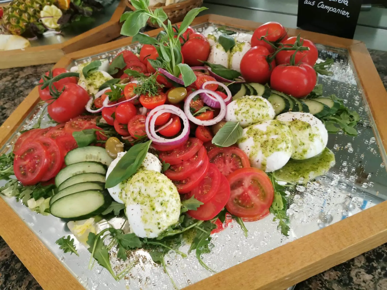 Mozzarellakugeln mit Kräuteröl neben Tomatenscheiben und Gurken auf verspiegeltem Tablett