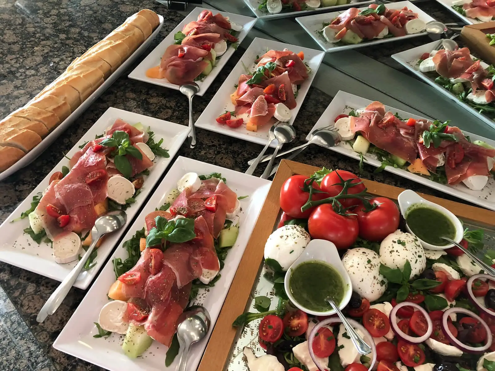 Parmaschinken mit Mozzarella und Melone auf weißen Platten, daneben aufgeschnittenes Baguette
