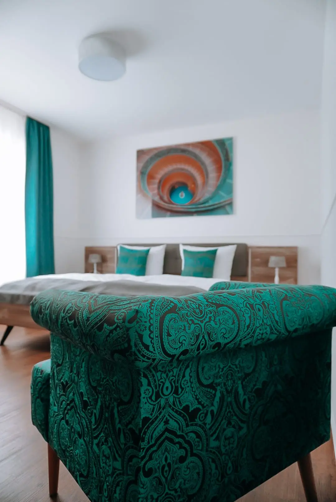 Grüner Ornamentsessel vor Doppelbett mit türkisen Kissen und Wendeltreppenbild an der Wand